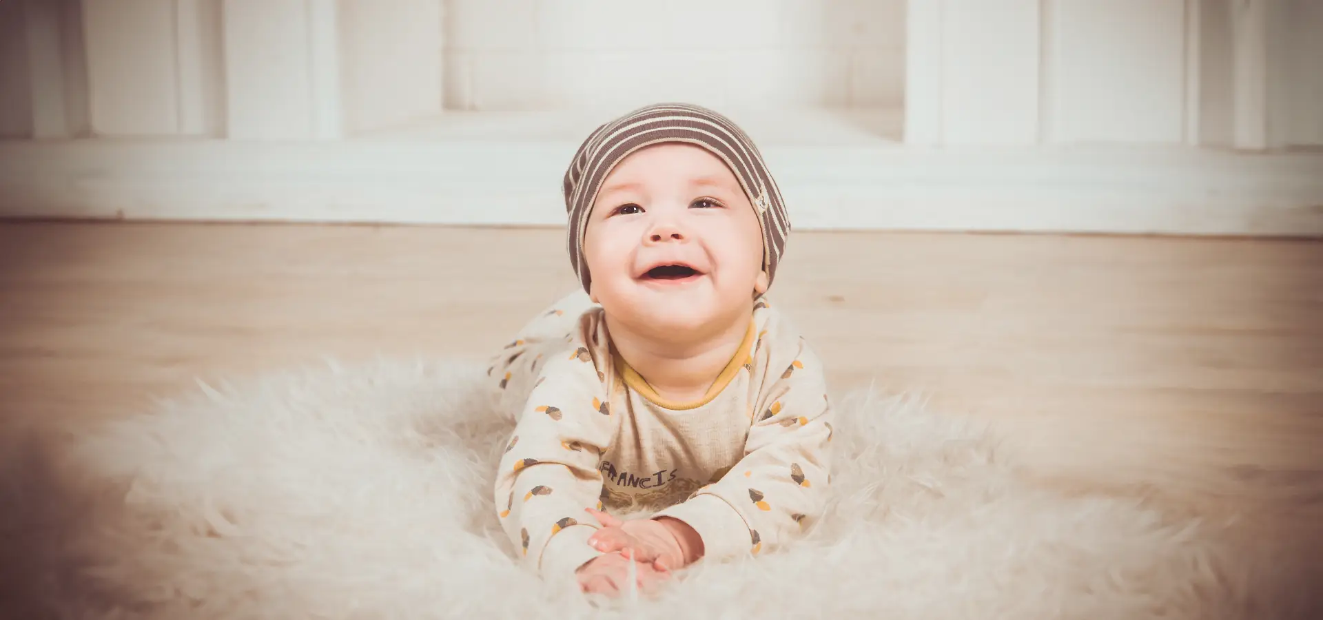Lachendes Baby mit Mütze auf weißem Fell liegend.