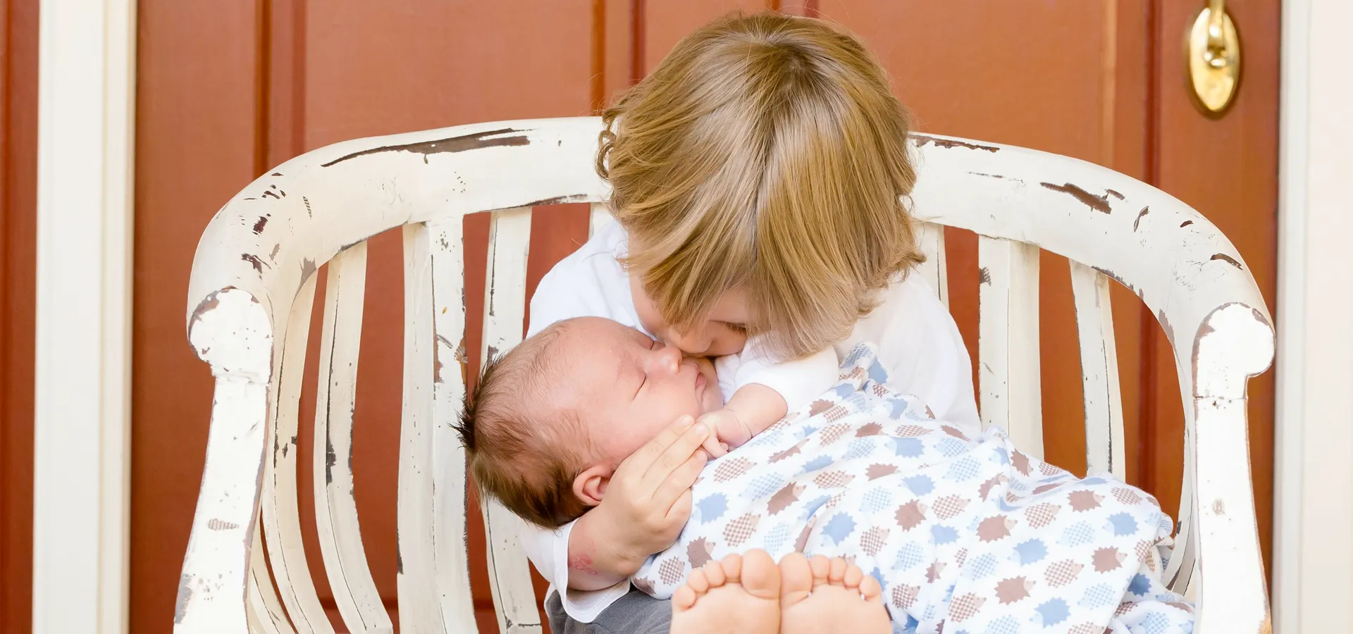 Kind gibt Neugeborenem auf Bank einen Kuss.