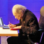 Ältere Person beim Schreiben an einem Tisch mit Laptop und Smartphone.