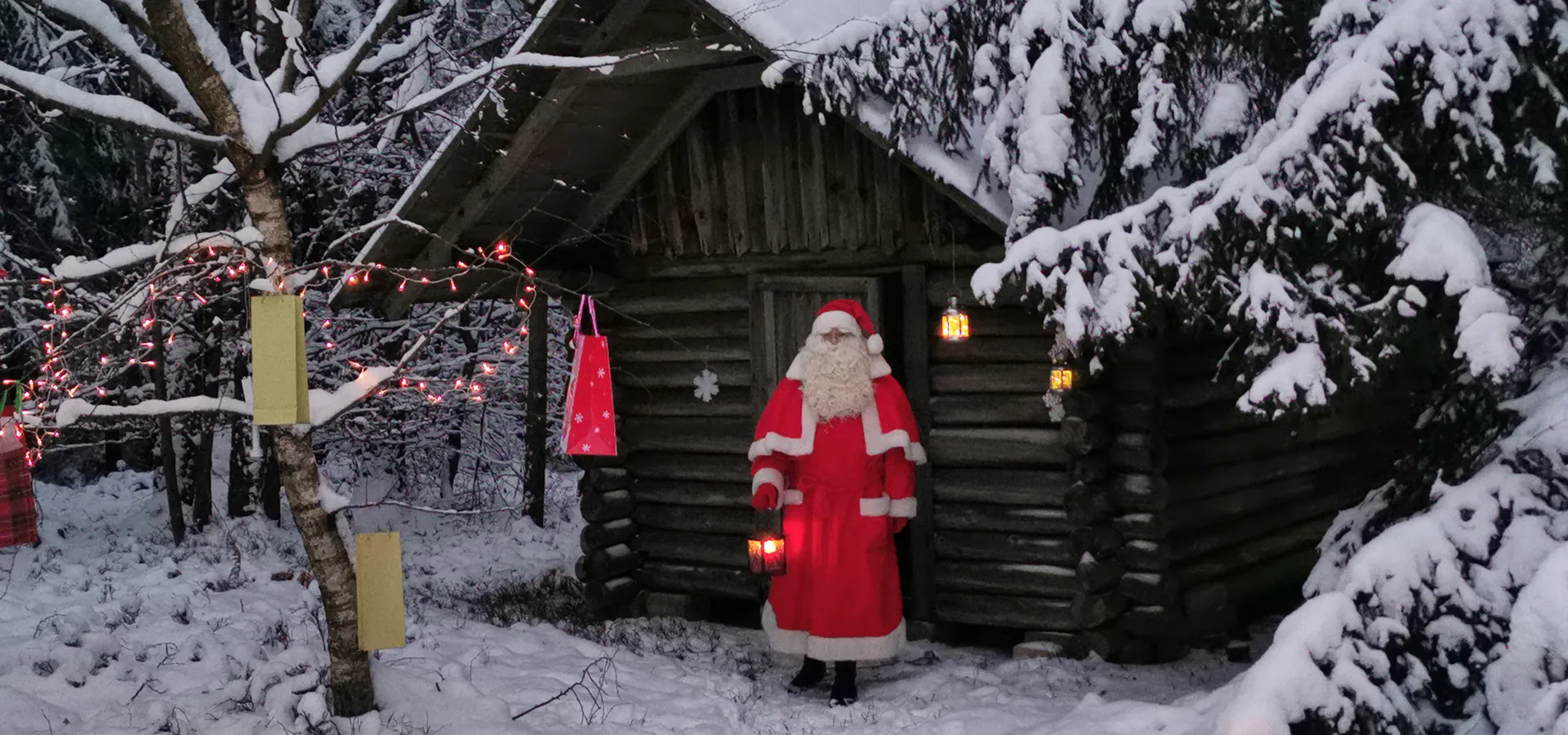 Weihnachtsmann vor einer Holzhütte im verschneiten Wald mit Lichterketten.
